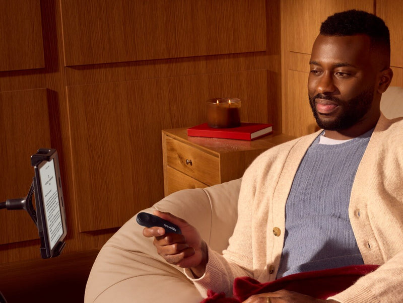 Un hombre, sentado cómodamente, sostiene Kobo Remote en su mano derecha mientras mira un eReader Kobo sobre un soporte flexible.