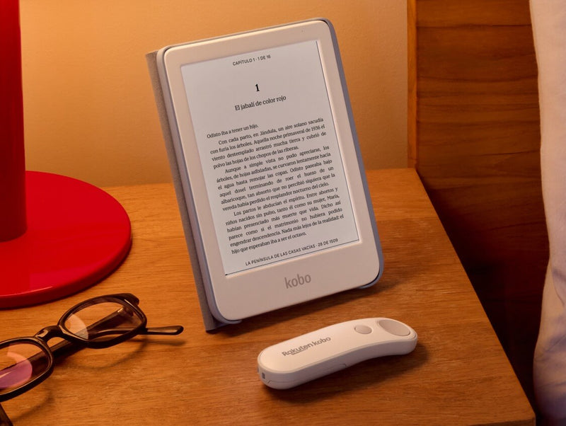 Un Kobo Clara Colour blanco está colocado sobre una mesa en su funda SleepCover con soporte incorporado, junto a un Kobo Remote blanco y un par de gafas.