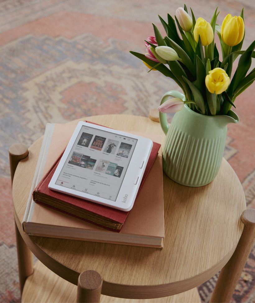 Un eReader Kobo Libra Colour apoyado sobre dos libros de tapa dura, colocados en una mesa de madera junto a un jarrón verde con flores.